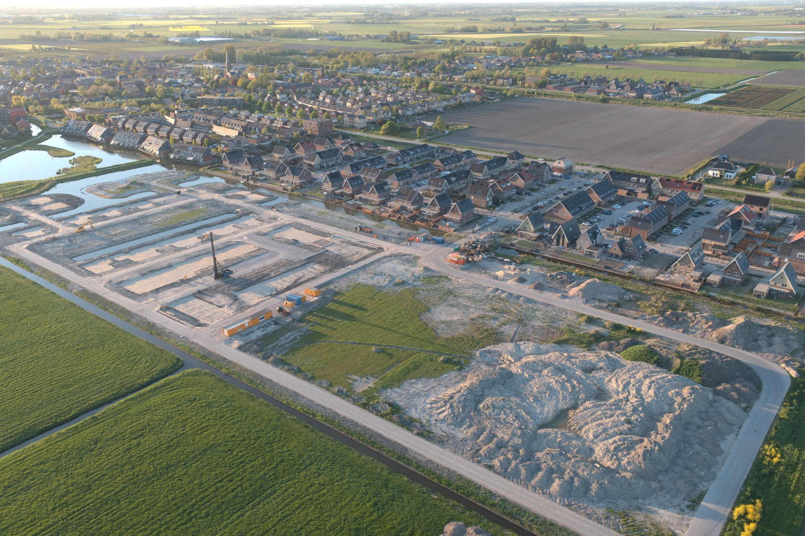 Luchtfoto van een nieuwe woonwijk in aanbouw aan het water, met de dorpskern op de achtergrond.