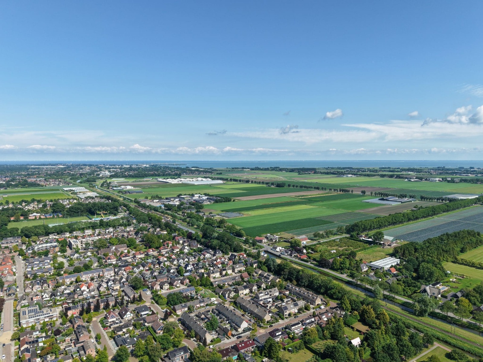 Luchtfoto van Westfriesland: akkers, een dorpsrand en de kust van het IJsselmeer in de verte.